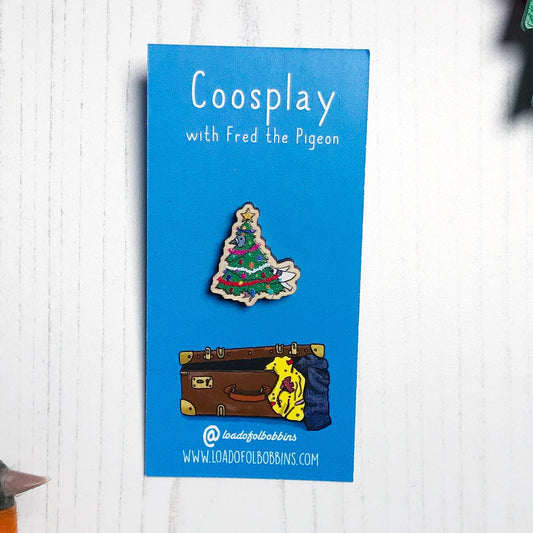 A laser cut wooden mini lapel pin showing an original illustration by Loadofolbobbins of a pigeon dressed as a Christmas Tree, complete with tinsel, decorations and star. The style of the illustration is simplified with flat colours and a bold black outline. The pin is mounted on a blue backing card that says Coosplay with Fred the pigeon.