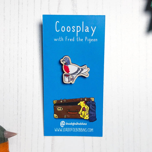 A laser cut wooden mini lapel pin showing an original illustration by Loadofolbobbins of a pigeon painting it's chest red to look like a robin. The style of the illustration is simplified with flat colours and a bold black outline. The pin is mounted on a blue backing card that says Coosplay with Fred the pigeon.