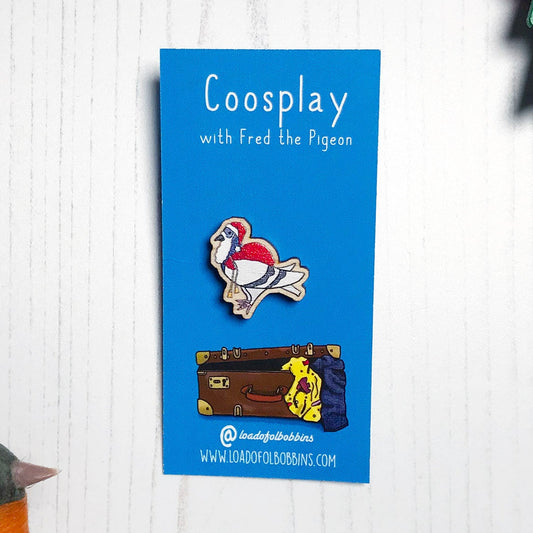 A laser cut wooden mini lapel pin showing an original illustration by Loadofolbobbins of a pigeon painting dressed as Santa Claus with a present sack on it's back. The style of the illustration is simplified with flat colours and a bold black outline. The pin is mounted on a blue backing card that says Coosplay with Fred the pigeon.