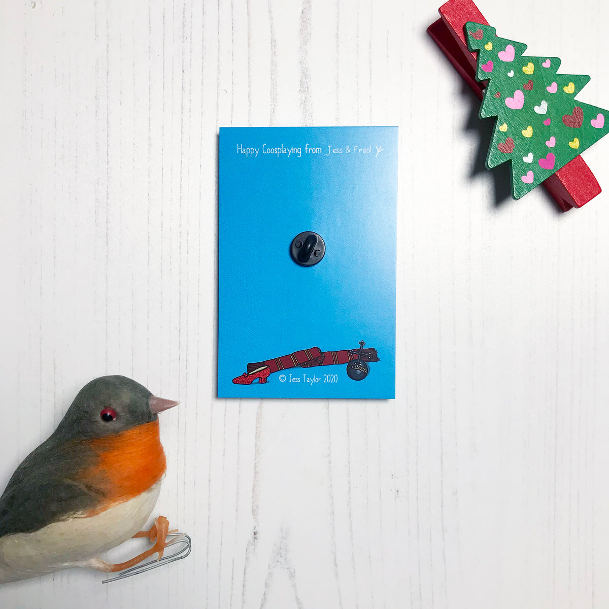 The back of a wooden lapel pin mounted on a blue backing card. You can see a black rubber clutch and text that says Happy coosplaying from Jess and Fred. Copyright Jess Taylor 2020.