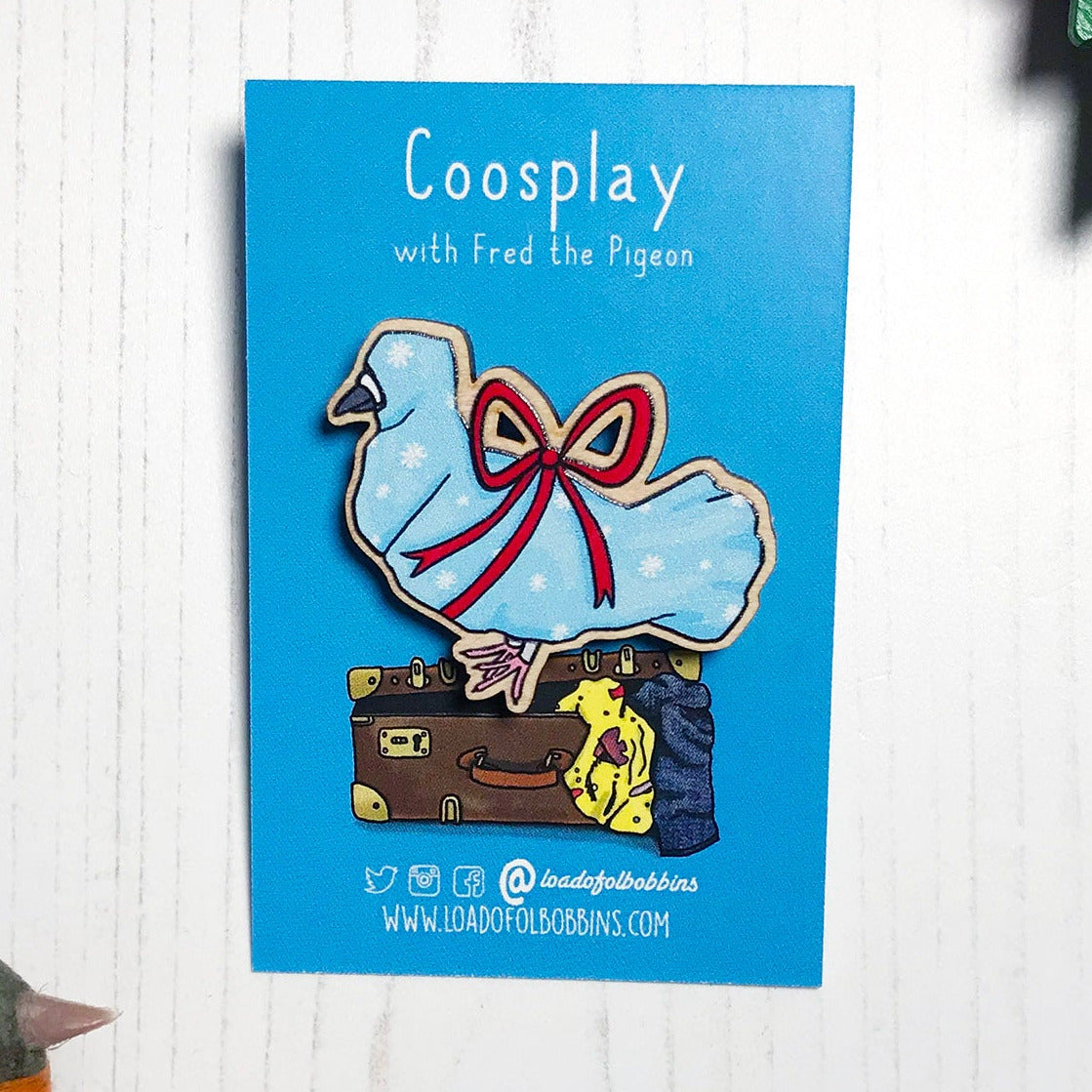 A laser cut wooden lapel pin showing an original illustration by Loadofolbobbins of a pigeon wrapped up like a Christmas present with a big red ribbon,it's beak and feet are peeking out of the wrapping paper. The style of the illustration is simplified with flat colours and a bold black outline. The pin is mounted on a blue backing card that says Coosplay with Fred the pigeon.