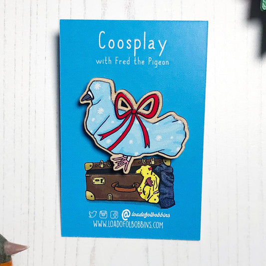 A laser cut wooden lapel pin showing an original illustration by Loadofolbobbins of a pigeon wrapped up like a Christmas present with a big red ribbon,it's beak and feet are peeking out of the wrapping paper. The style of the illustration is simplified with flat colours and a bold black outline. The pin is mounted on a blue backing card that says Coosplay with Fred the pigeon.