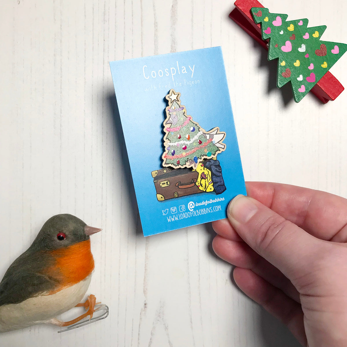 A laser cut wooden lapel pin showing an original illustration by Loadofolbobbins of a pigeon dressed as a Christmas Tree, complete with tinsel, decorations and star. The style of the illustration is simplified with flat colours and a bold black outline. The pin is mounted on a blue backing card that says Coosplay with Fred the pigeon. The pin is held at an angle showing of the spot gloss detail on the decorations and star.