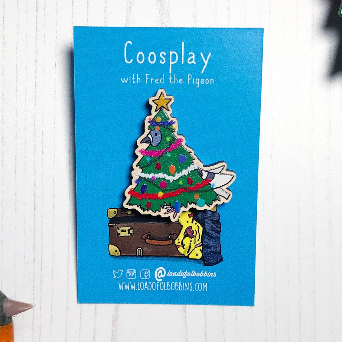 A laser cut wooden lapel pin showing an original illustration by Loadofolbobbins of a pigeon dressed as a Christmas Tree, complete with tinsel, decorations and star. The style of the illustration is simplified with flat colours and a bold black outline. The pin is mounted on a blue backing card that says Coosplay with Fred the pigeon.