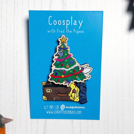A laser cut wooden lapel pin showing an original illustration by Loadofolbobbins of a pigeon dressed as a Christmas Tree, complete with tinsel, decorations and star. The style of the illustration is simplified with flat colours and a bold black outline. The pin is mounted on a blue backing card that says Coosplay with Fred the pigeon.