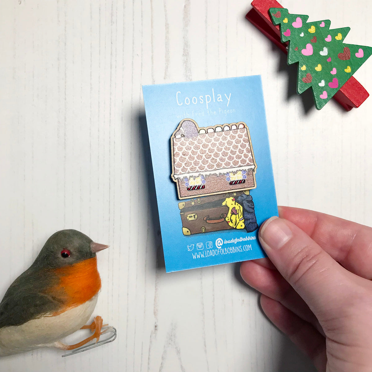 A laser cut wooden lapel pin showing an original illustration by Loadofolbobbins of a pigeon with it's head popping out of a gingerbread house nibbling on some jelly sweets.. The style of the illustration is simplified with flat colours and a bold black outline. The pin is mounted on a blue backing card that says Coosplay with Fred the pigeon. A hand is holding it at an angle to show light shining off spot gloss detail on the candy can and jelly tot details.