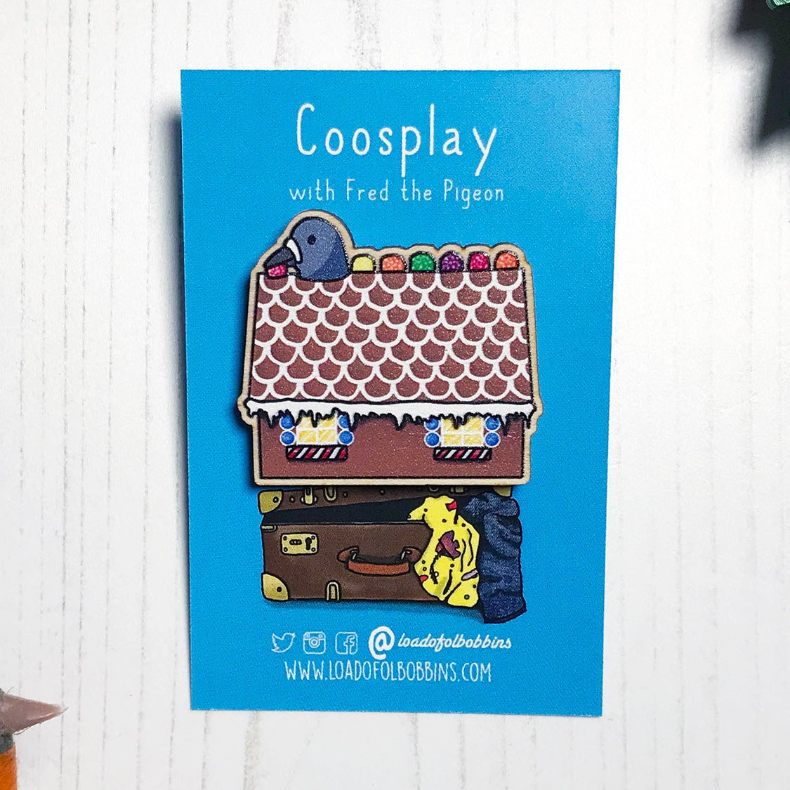 A laser cut wooden lapel pin showing an original illustration by Loadofolbobbins of a pigeon with it's head popping out of a gingerbread house nibbling on some jelly sweets.. The style of the illustration is simplified with flat colours and a bold black outline. The pin is mounted on a blue backing card that says Coosplay with Fred the pigeon.