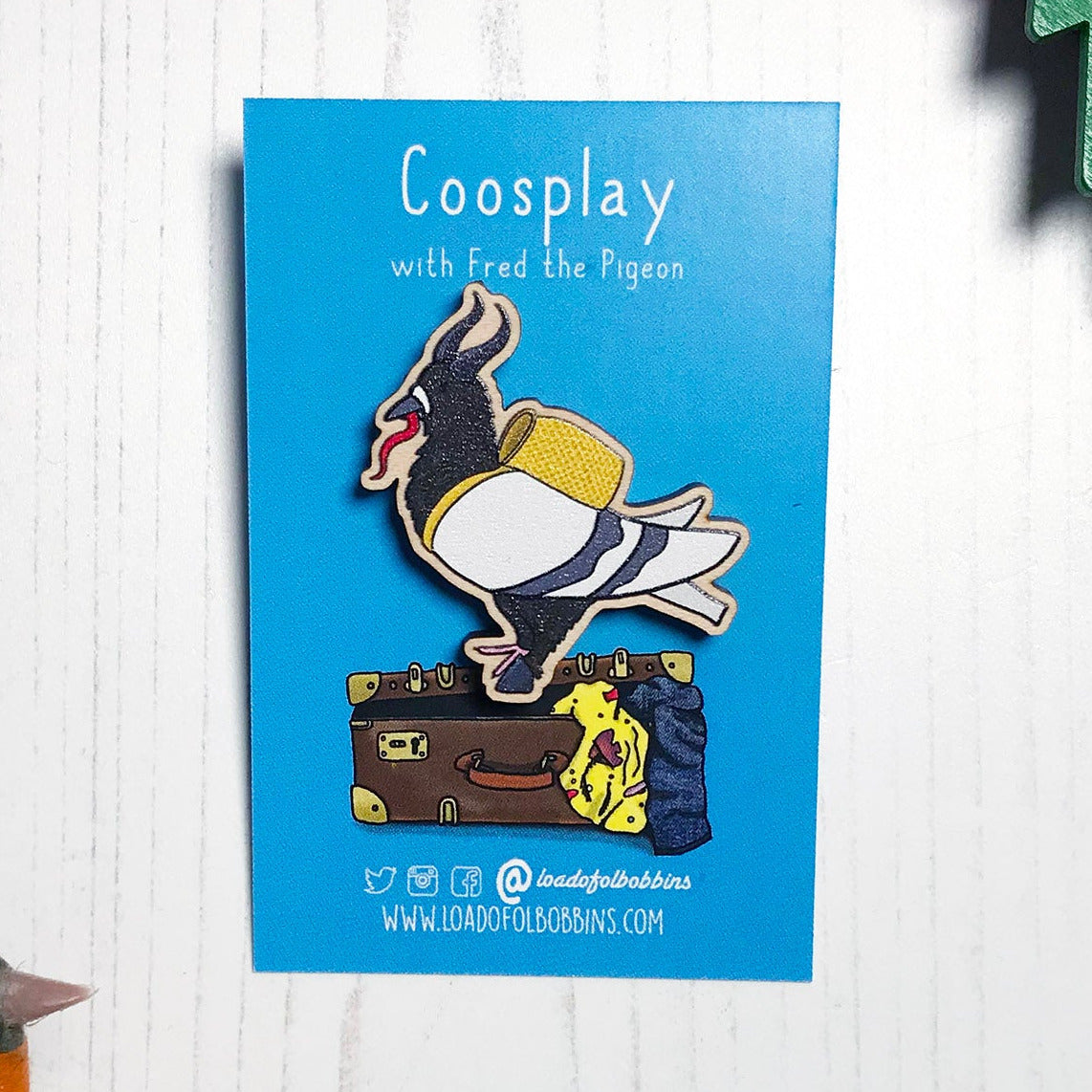 A laser cut wooden lapel pin showing an original illustration by Loadofolbobbins of a pigeon dressed as Krampus, with horns a cloven hoof and long tongue. The style of the illustration is simplified with flat colours and a bold black outline. The pin is mounted on a blue backing card that says Coosplay with Fred the pigeon.