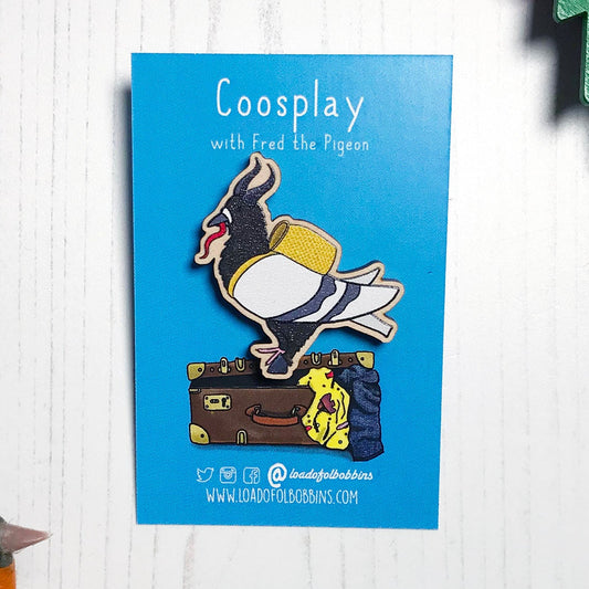 A laser cut wooden lapel pin showing an original illustration by Loadofolbobbins of a pigeon dressed as Krampus, with horns a cloven hoof and long tongue. The style of the illustration is simplified with flat colours and a bold black outline. The pin is mounted on a blue backing card that says Coosplay with Fred the pigeon.