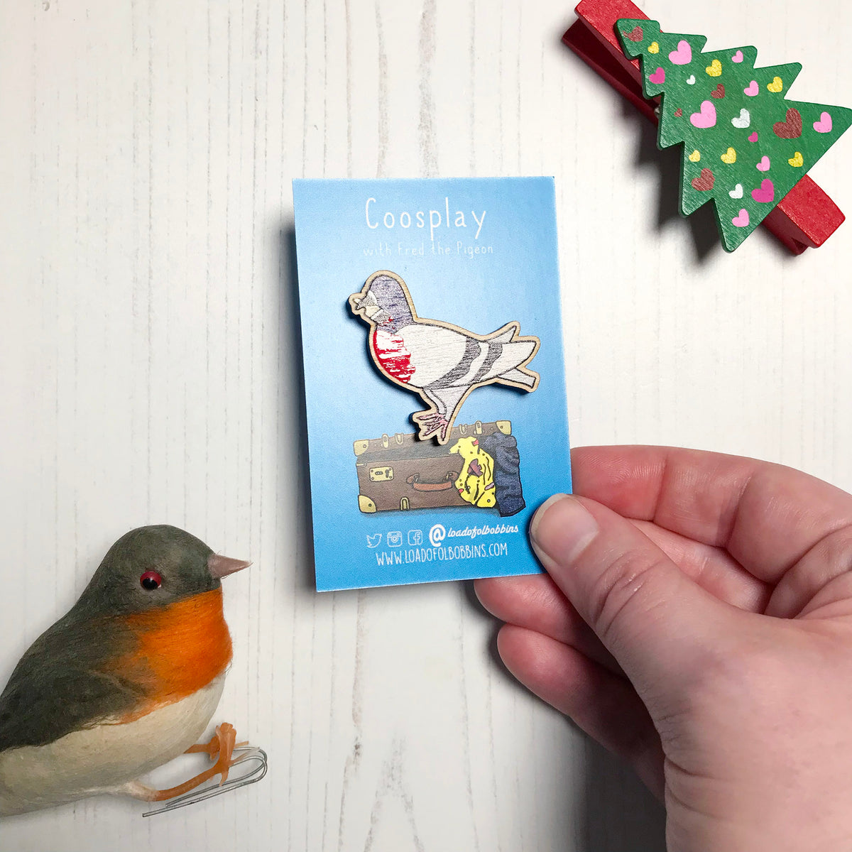 A laser cut wooden lapel pin showing an original illustration by Loadofolbobbins of a pigeon painting it's chest red to look like a robin. The style of the illustration is simplified with flat colours and a bold black outline. The pin is mounted on a blue backing card that says Coosplay with Fred the pigeon. The pin is held at an angle to show off the spot gloss detail on the red painted chest.