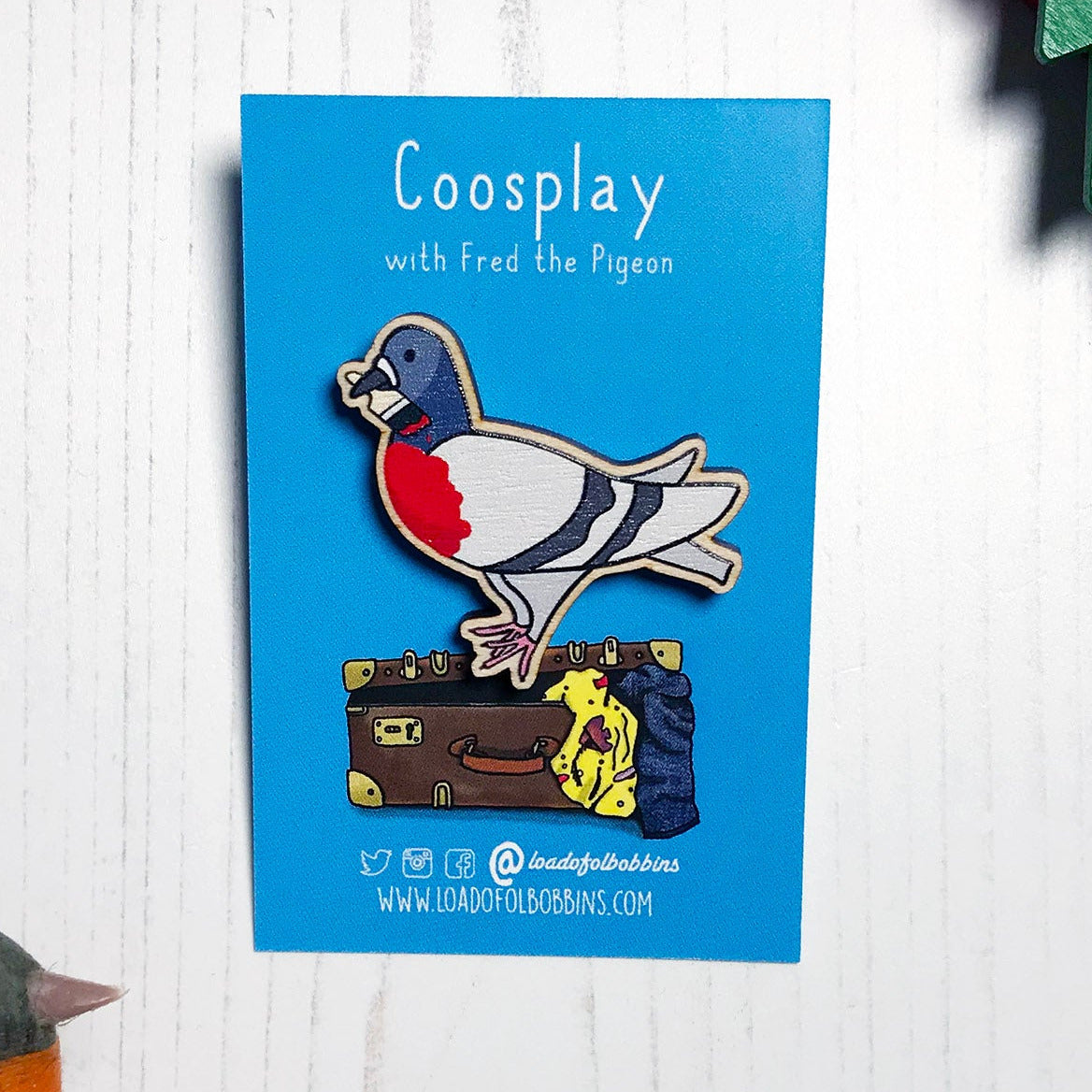 A laser cut wooden lapel pin showing an original illustration by Loadofolbobbins of a pigeon painting it's chest red to look like a robin. The style of the illustration is simplified with flat colours and a bold black outline. The pin is mounted on a blue backing card that says Coosplay with Fred the pigeon.