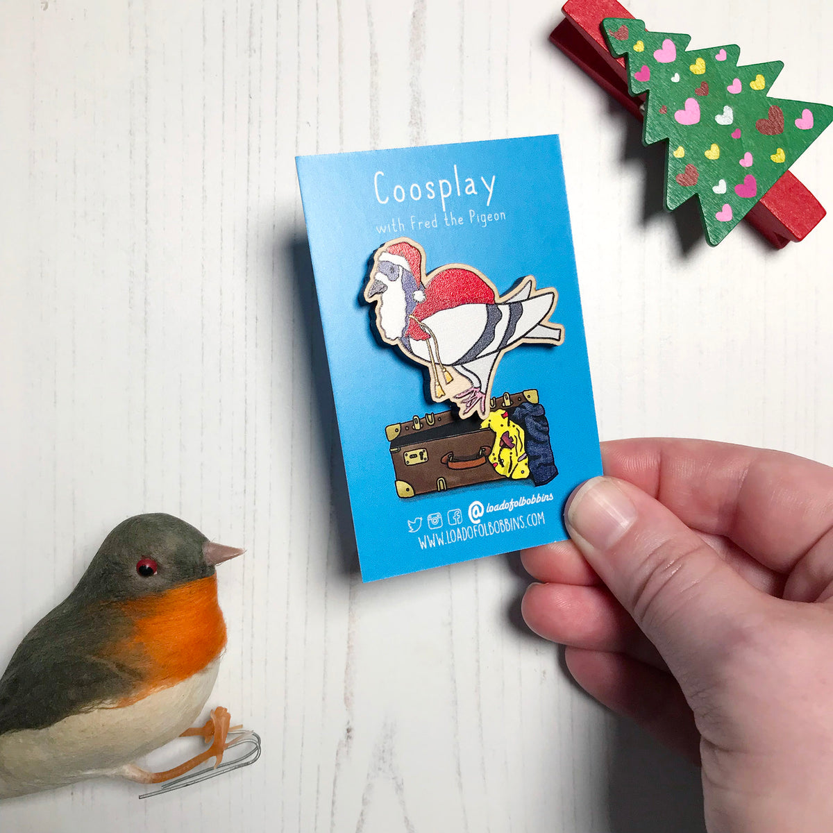 A laser cut wooden lapel pin showing an original illustration by Loadofolbobbins of a pigeon painting dressed as Santa Claus with a present sack on it's back. The style of the illustration is simplified with flat colours and a bold black outline. The pin is mounted on a blue backing card that says Coosplay with Fred the pigeon. The pin is shown at an angle.