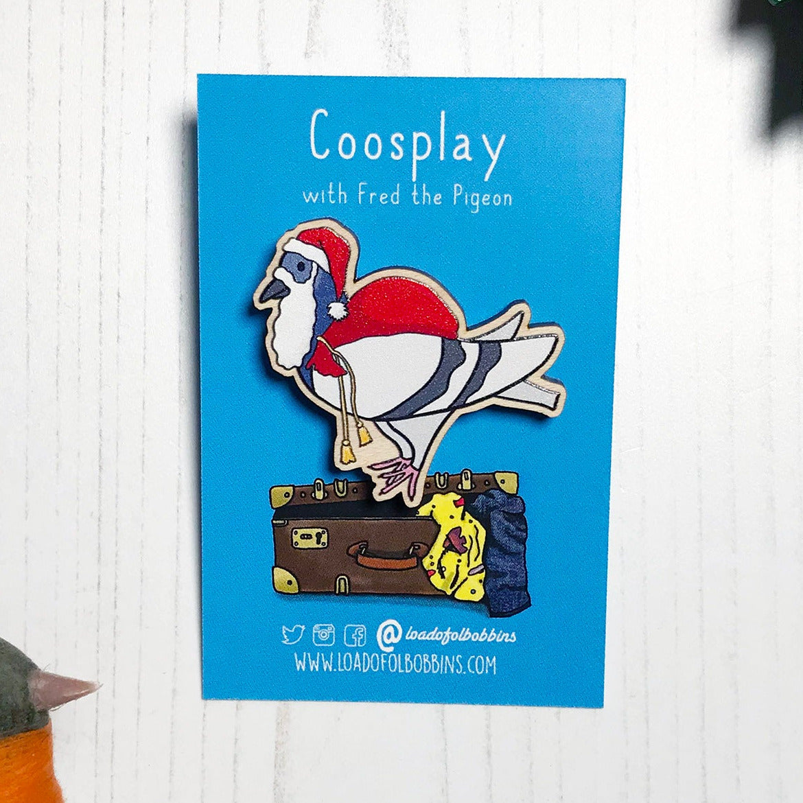 A laser cut wooden lapel pin showing an original illustration by Loadofolbobbins of a pigeon painting dressed as Santa Claus with a present sack on it's back. The style of the illustration is simplified with flat colours and a bold black outline. The pin is mounted on a blue backing card that says Coosplay with Fred the pigeon.