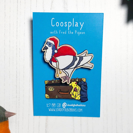 A laser cut wooden lapel pin showing an original illustration by Loadofolbobbins of a pigeon painting dressed as Santa Claus with a present sack on it's back. The style of the illustration is simplified with flat colours and a bold black outline. The pin is mounted on a blue backing card that says Coosplay with Fred the pigeon.