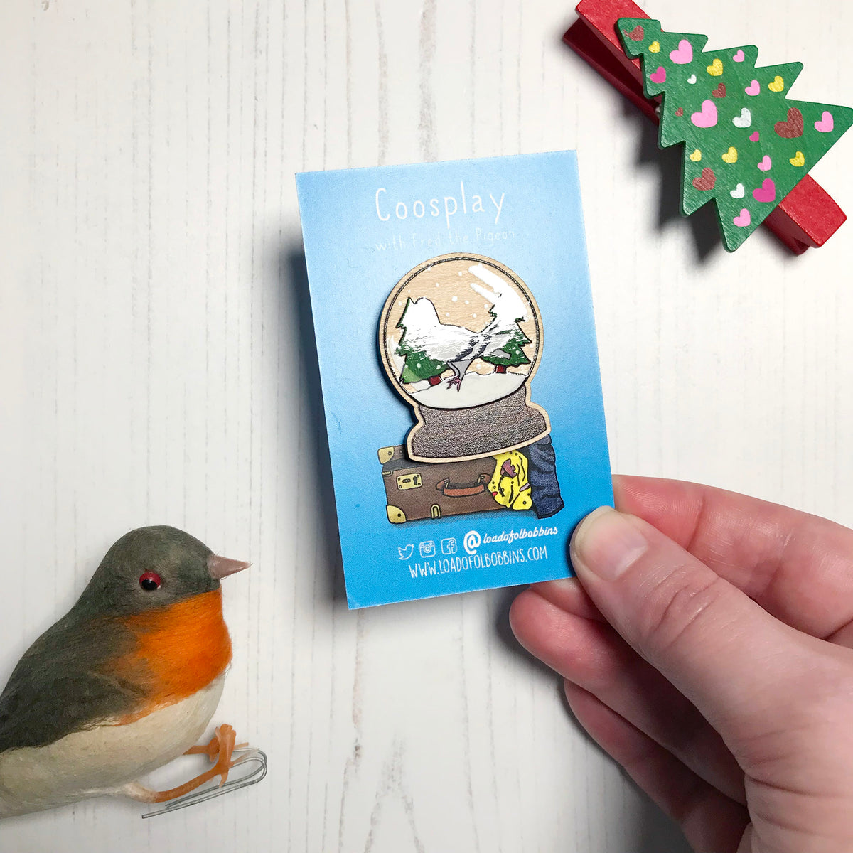 A laser cut wooden lapel pin showing an original illustration by Loadofolbobbins of a pigeon in a snow globe, standing on snow and next to two trees. The style of the illustration is simplified with flat colours and a bold black outline. The pin is mounted on a blue backing card that says Coosplay with Fred the pigeon. It's held by a hand at a slight angle highlighting the spot gloss detail on the pigeon and trees.