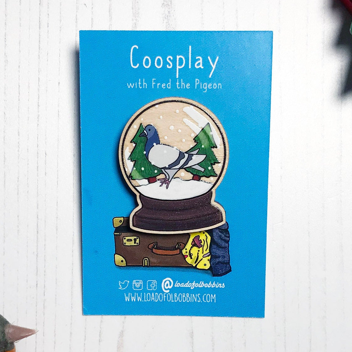 A laser cut wooden lapel pin showing an original illustration by Loadofolbobbins of a pigeon in a snow globe, standing on snow and next to two trees. The style of the illustration is simplified with flat colours and a bold black outline. The pin is mounted on a blue backing card that says Coosplay with Fred the pigeon.