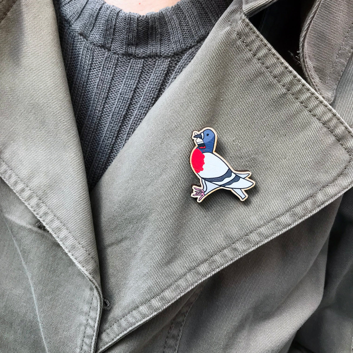 A laser cut wooden lapel pin showing an original illustration by Loadofolbobbins of a pigeon painting it's chest red to look like a robin. The style of the illustration is simplified with flat colours and a bold black outline. The pin is shown worn on the lapel of a jacket.