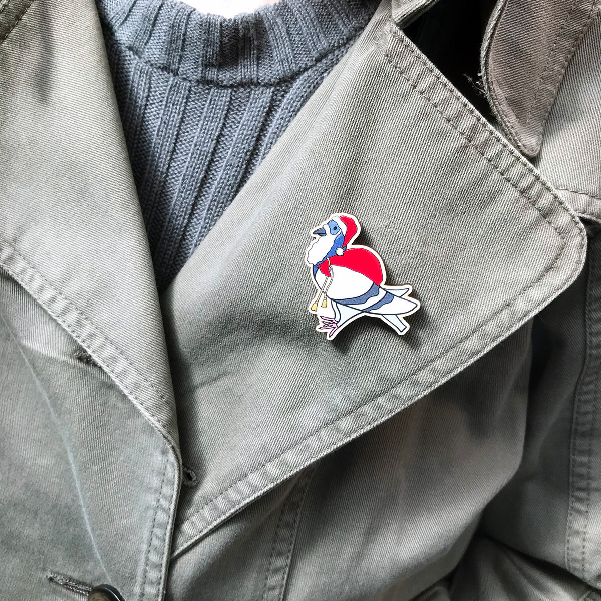 A laser cut wooden lapel pin showing an original illustration by Loadofolbobbins of a pigeon painting dressed as Santa Claus with a present sack on it's back. The style of the illustration is simplified with flat colours and a bold black outline. The pin is shown on the lapel of a jacket.