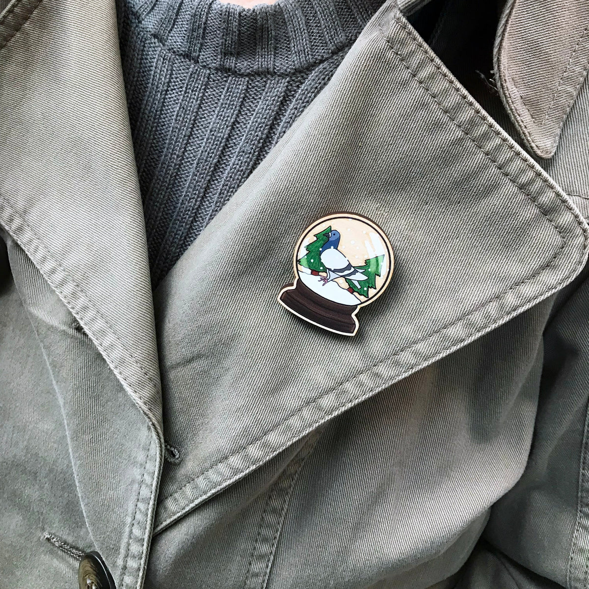 A laser cut wooden lapel pin showing an original illustration by Loadofolbobbins of a pigeon in a snow globe, standing on snow and next to two trees. The style of the illustration is simplified with flat colours and a bold black outline. The pin is shown worn on the lapel of a jacket.