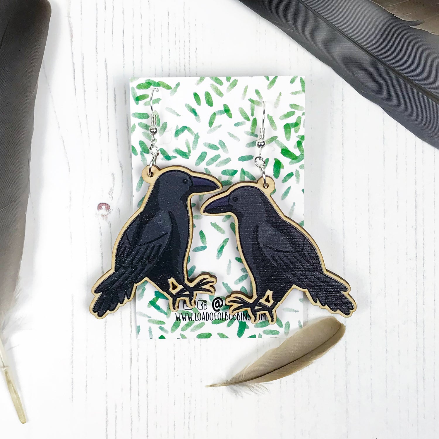 A pair of laser cut wooden dangle earrings showing an illustration by Loadofolbobbins of a crow. The earrings are mounted on a backing card with a repeat abstract watercolour leaf pattern.