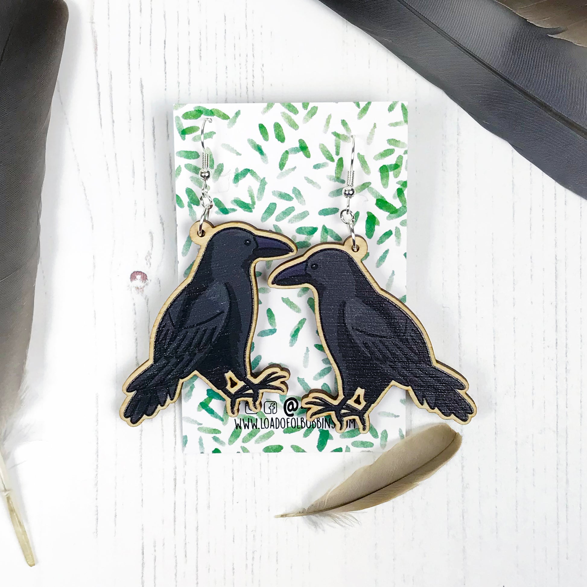 A pair of laser cut wooden dangle earrings showing an illustration by Loadofolbobbins of a crow. The earrings are mounted on a backing card with a repeat abstract watercolour leaf pattern.