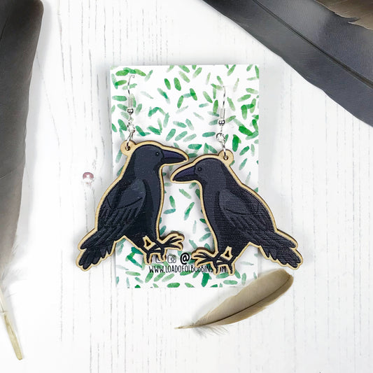 A pair of laser cut wooden dangle earrings showing an illustration by Loadofolbobbins of a crow. The earrings are mounted on a backing card with a repeat abstract watercolour leaf pattern.