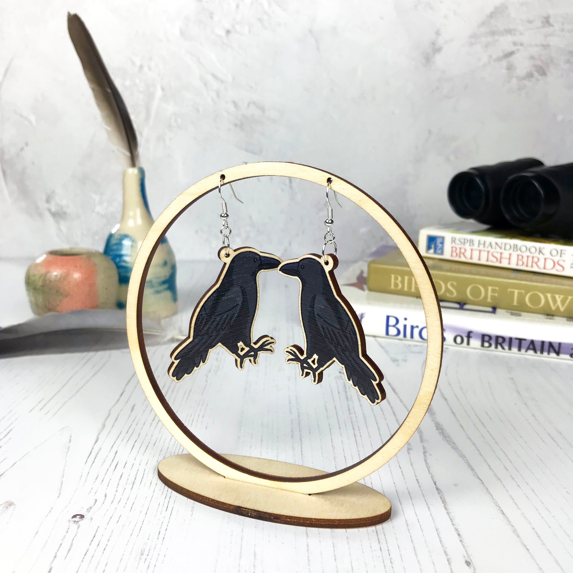 A pair of laser cut wooden dangle earrings showing an illustration of a crow bird by loadofolbobbins. The earrings are hanging from a wooden circular ring display. 