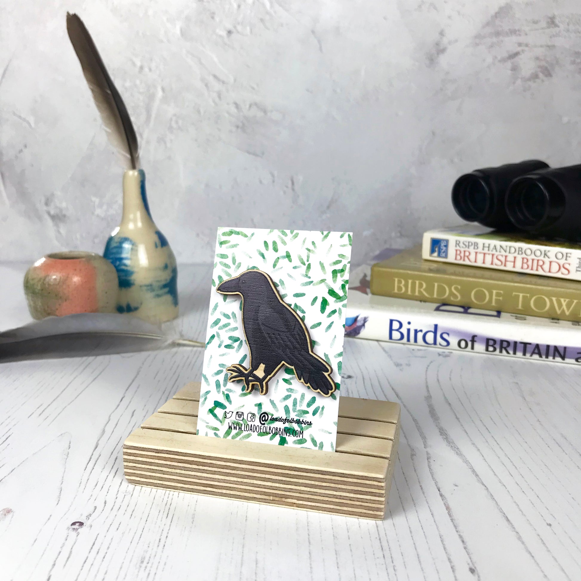 A laser cut wooden brooch showing an illustration of a crow bird by loadofolbobbins. The brooch is mounted on a backing card with a repeat abstract watercolour leaf pattern. They are standing in a wooden stand and in the background you can see a pile of bird watching books and a pair of binoculars.