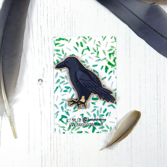 A laser cut wooden brooch showing an illustration by Loadofolbobbins of a crow. The brooch is mounted on a backing card with a repeat abstract watercolour leaf pattern.