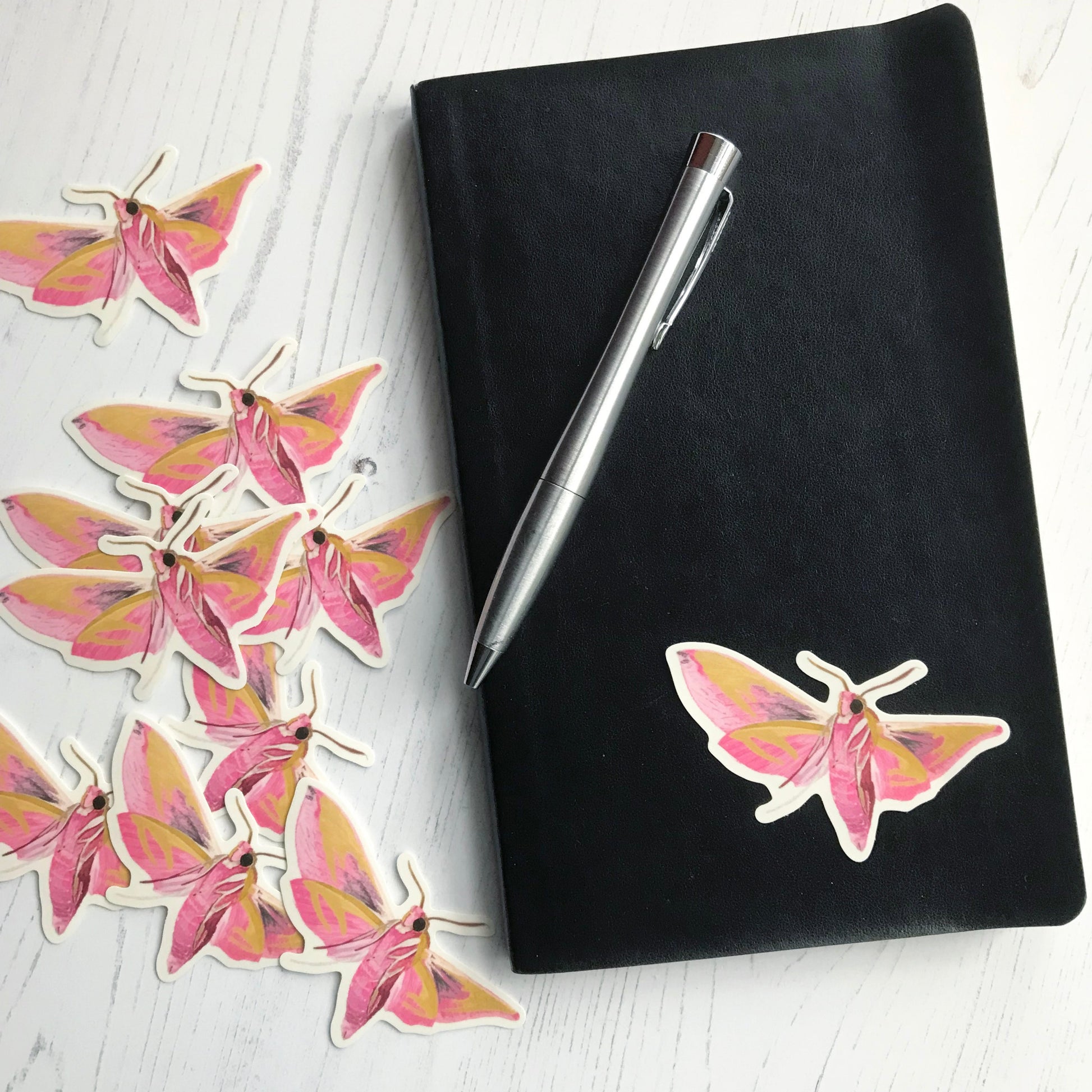 A elephant hawk-moth sticker created from an original digital illustration by Loadofolbobbins. The moth is shown mid flight. It's striking thick pink segmented lower abdomen and fluffy upper body can be seen with three pale legs either side. It's four wings are spread wide, with incredible warm pink colouring and an almost mustard gold area of markings at the top edge of each wing. The sticker is on a black notebook with a pen resting on it.