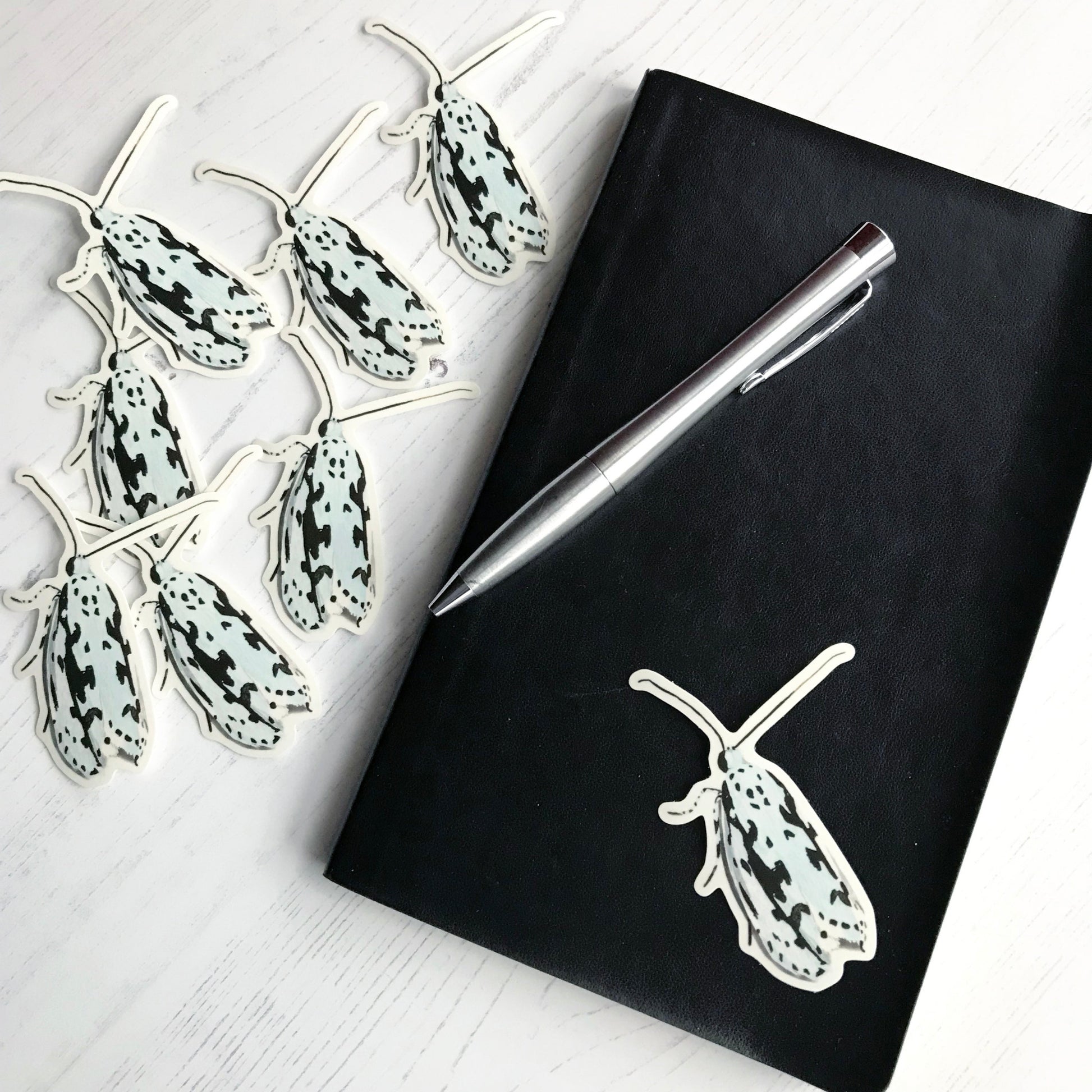 A ethmia pusiela moth sticker created from an original digital illustration by Loadofolbobbins. A small delicate moth with it's wings closed behind itself. It is predominantly white with distinctive black markings across it wings and legs, very like the Rorschach test. The sticker is on a black notebook with a pen resting on it.