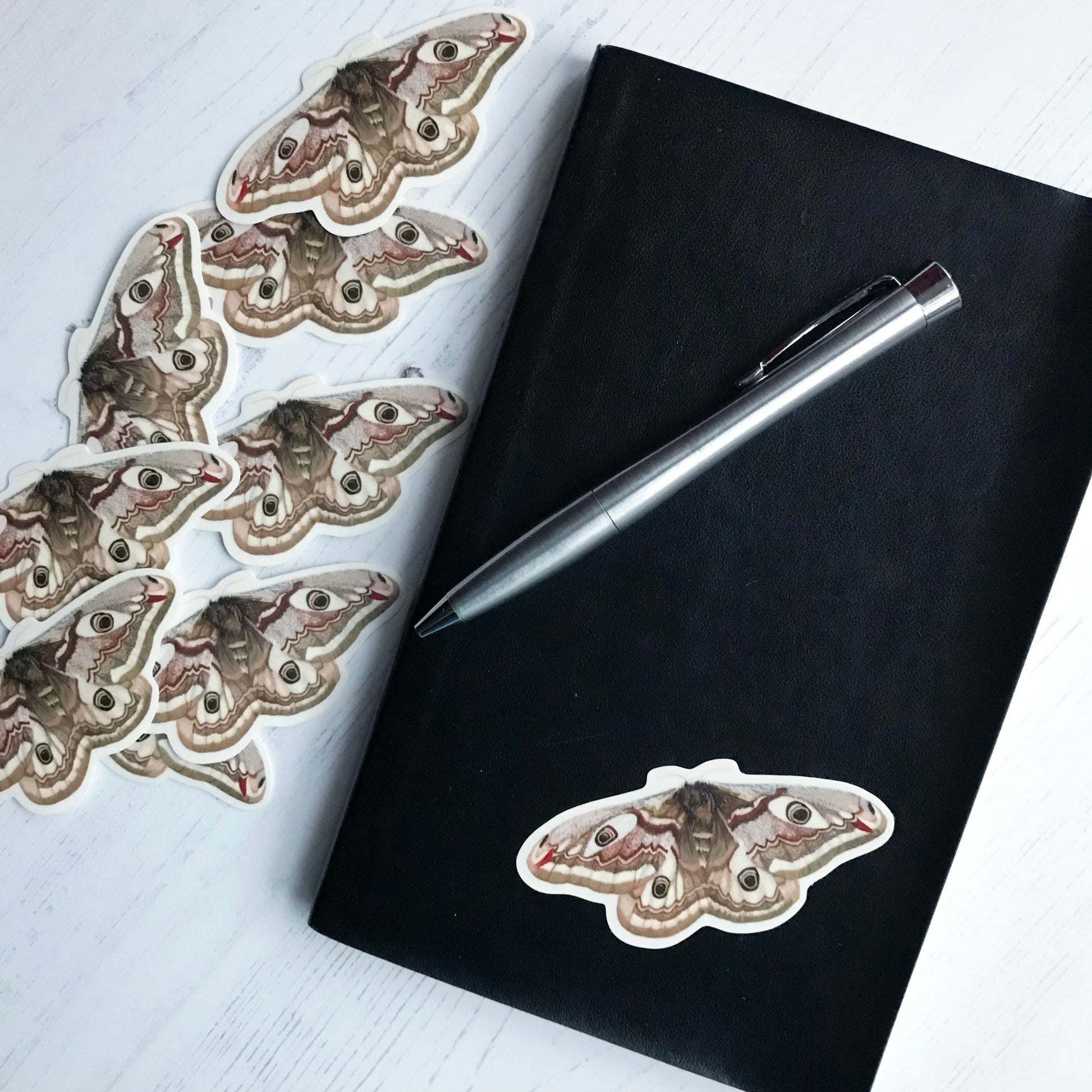 A female emperor moth sticker created from an original digital illustration by Loadofolbobbins. The moth's small creamy coloured antennae lead to a dark brown fluffy body with a white stripe at the top. It's a large moth with intricately patterned wings. At the base of each wing is a mid brown stripe and a creamy white stripe above it, above this is an area of dark brown made up of tiny dots almost like a pointillist painting. On each wing is a peacock like eye spot in a cream area. 