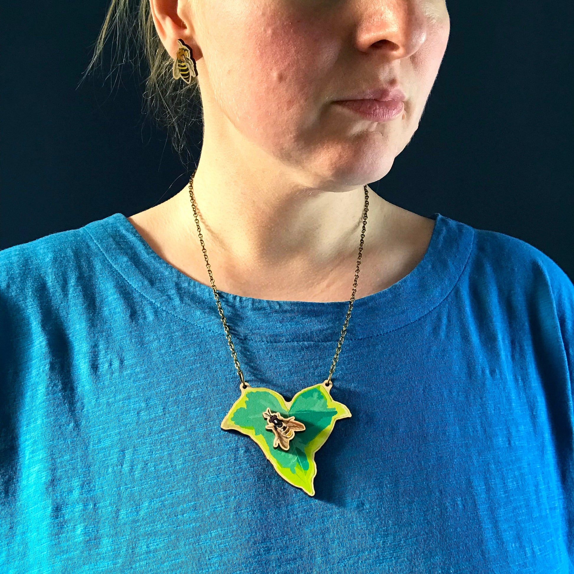 Golden Hoverfly and Ivy necklace by Loadofolbobbins. A laser cut wooden necklace that shows a small ivy leaf with a golden hoverfly on. The necklace hangs on an antique bronze chain. It is being worn by me a white woman wearing a light blue top. You can just see the bottom of my face and neck.
