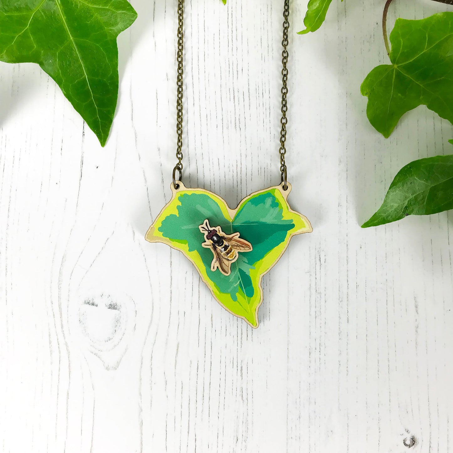 A laser cut wooden statement necklace showing an illustration of an variegated ivy leaf and a golden hoverfly by loadofolbobbins. The hoverfly stands proud of the necklace in a 3D relief effect.  The wooden necklace is hanging from an antique bronze cable chain.