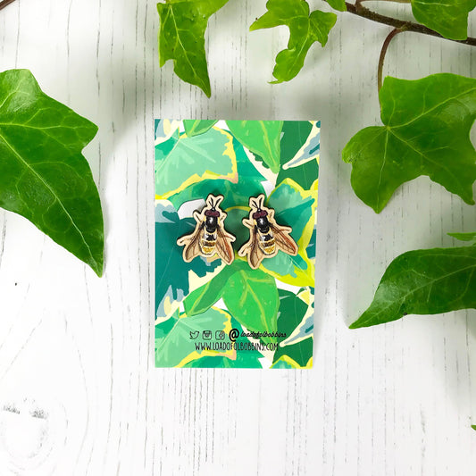 A pair of laser cut matching wooden stud earrings showing an illustration by Loadofolbobbins of a golden hoverfly. The earrings are mounted on a backing card with a repeat ivy leaf pattern.