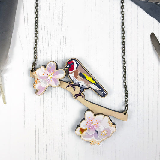 A laser cut wooden charm necklace showing an illustration of a goldfinch bird perching on a branch with cherry blossoms by loadofolbobbins. The cherry blossom flowers stand proud of the necklace in a 3D relief effect.  The wooden necklace is hanging from an antique bronze cable chain.
