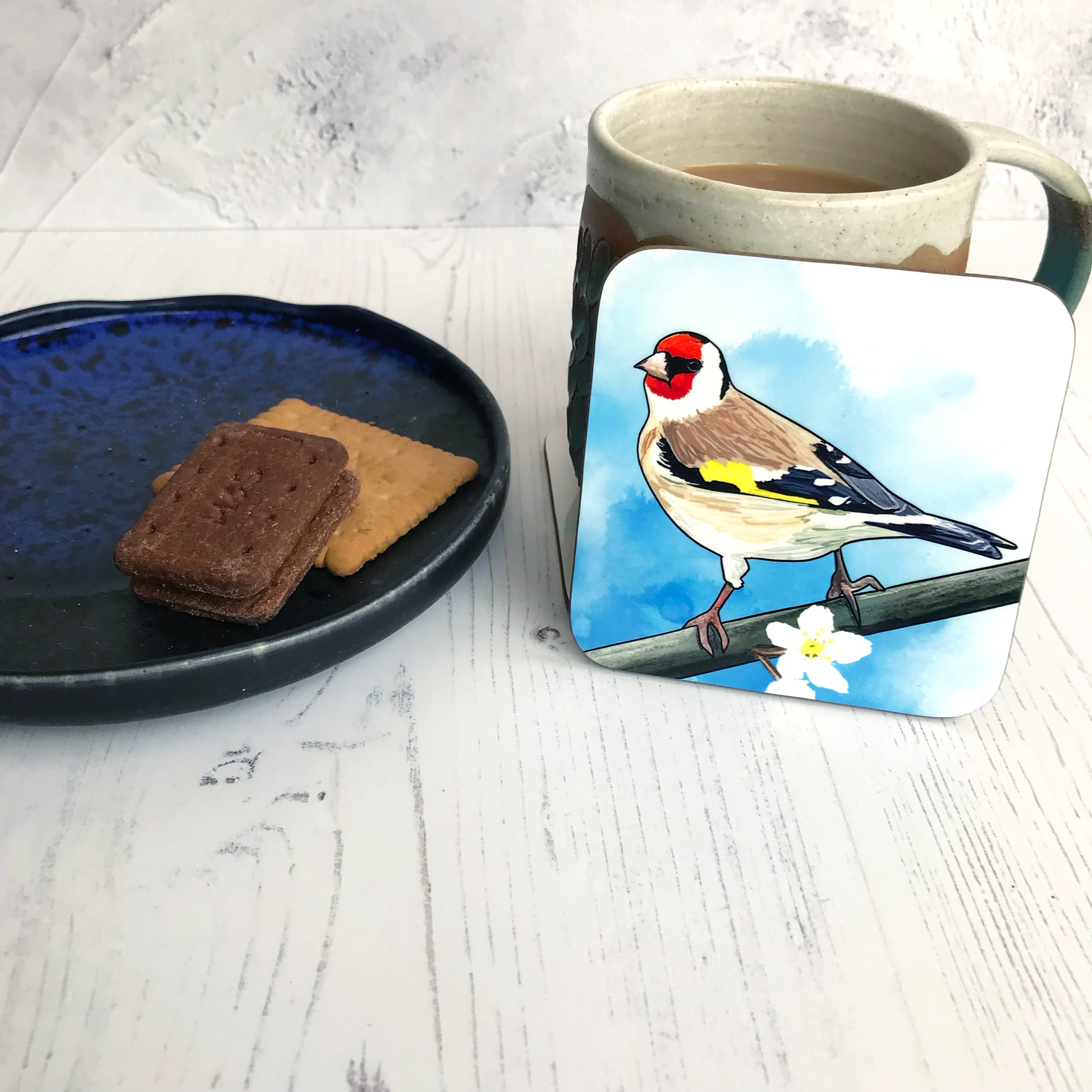 A photo of a round edged square coaster which has an original illustration by Loadofolbobbins showing a European goldfinch, back facing the viewer, head turned and looking out towards the left. It has a bright red face with black eye band and a creamy white underbelly with brown wings which are black towards the end with flashes of white, also a bright yellow flash of feathers on their wing. It is perching on a branch with white tree blossoms.