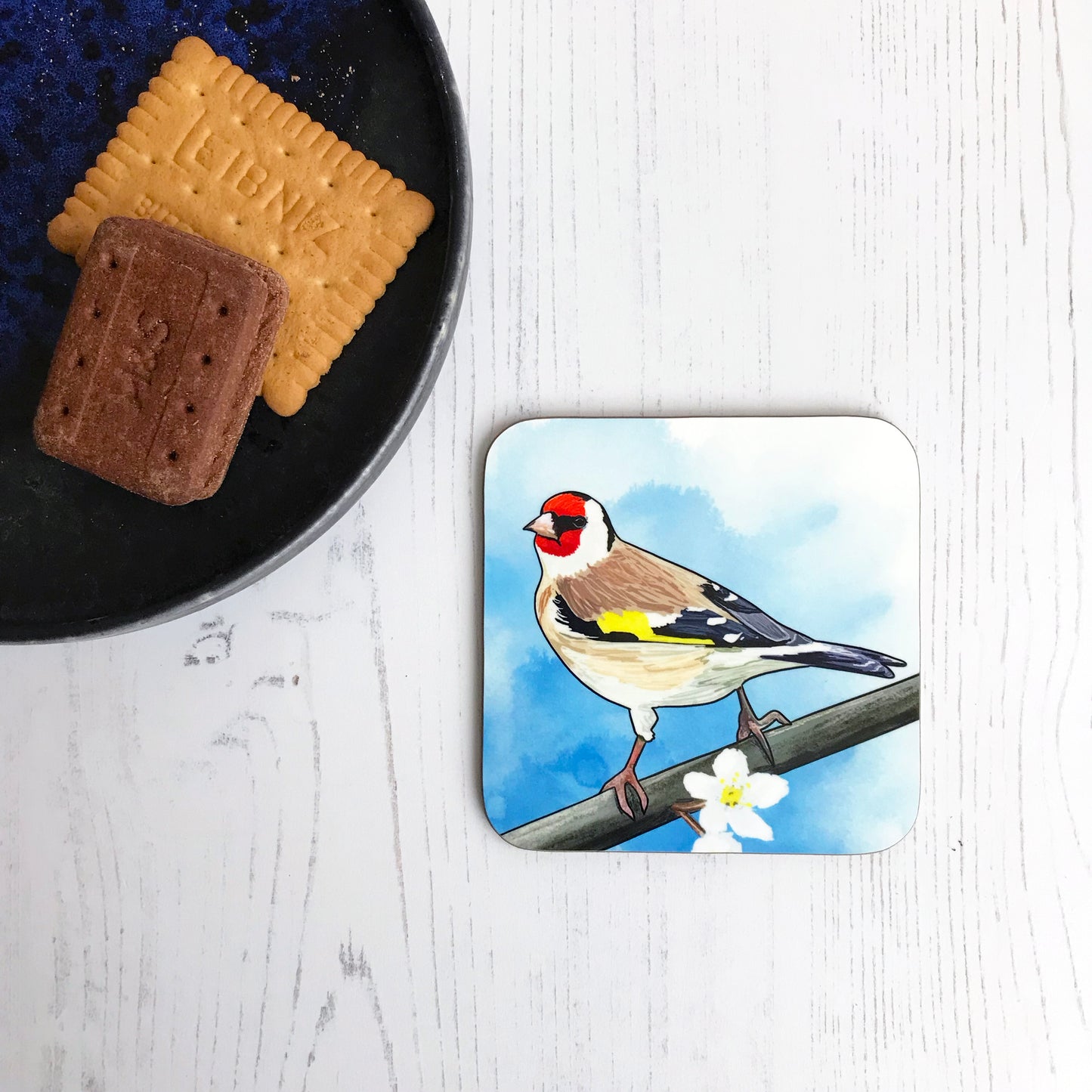 A photo of a round edged square coaster which has an original illustration by Loadofolbobbins showing a European goldfinch, back facing the viewer, head turned and looking out towards the left. It has a bright red face with black eye band and a creamy white underbelly with brown wings which are black towards the end with flashes of white, also a bright yellow flash of feathers on their wing. It is perching on a branch with white tree blossoms.