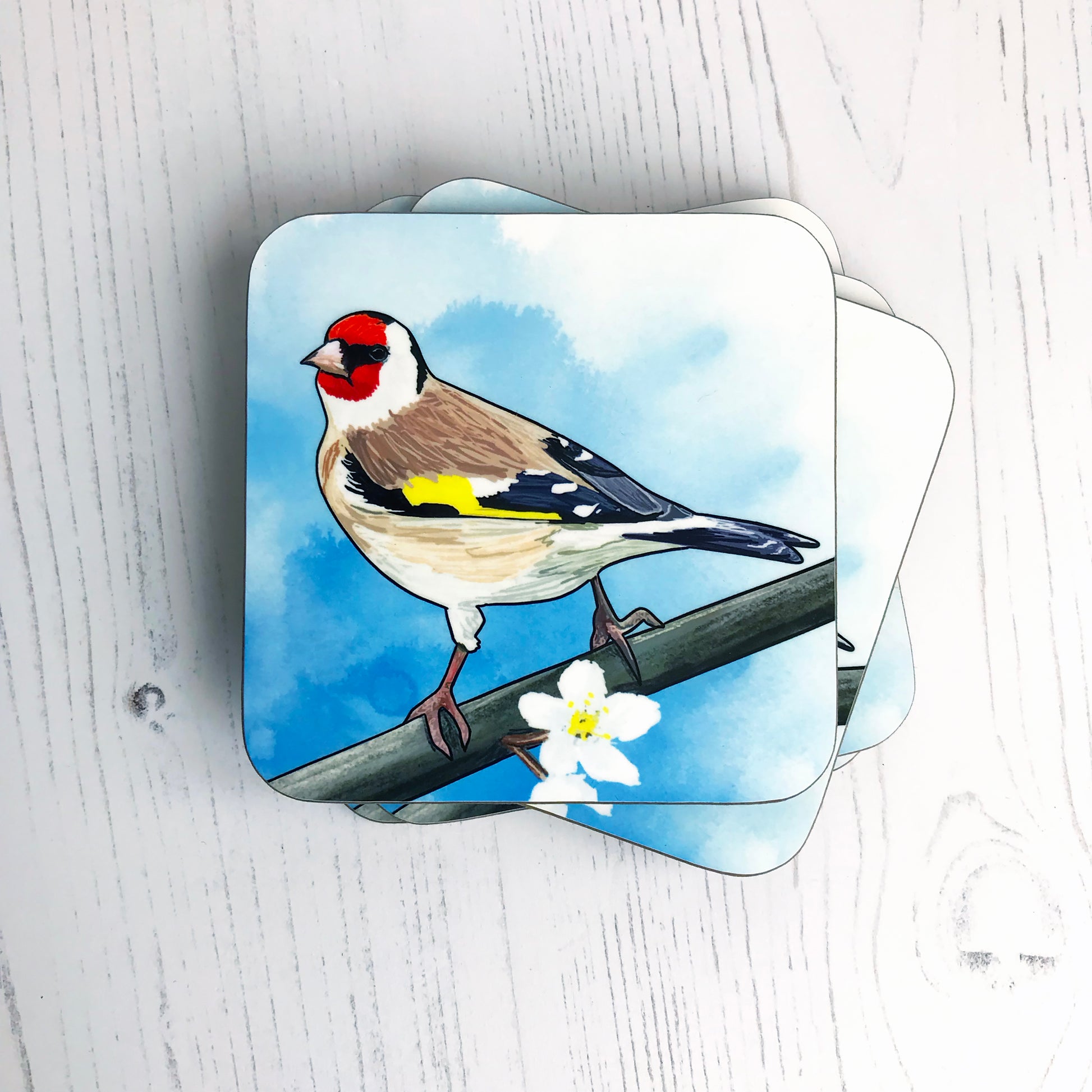 A photo of a pile of round edged square coasters which have an original illustration by Loadofolbobbins showing a European goldfinch, back facing the viewer, head turned and looking out towards the left. It has a bright red face with black eye band and a creamy white underbelly with brown wings which are black towards the end with flashes of white, also a bright yellow flash of feathers on their wing. It is perching on a branch with white tree blossoms.