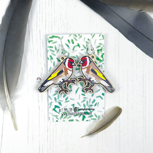 A pair of laser cut wooden dangle earrings showing an illustration by Loadofolbobbins of a goldfinch. The earrings are mounted on a backing card with a repeat abstract watercolour leaf pattern.