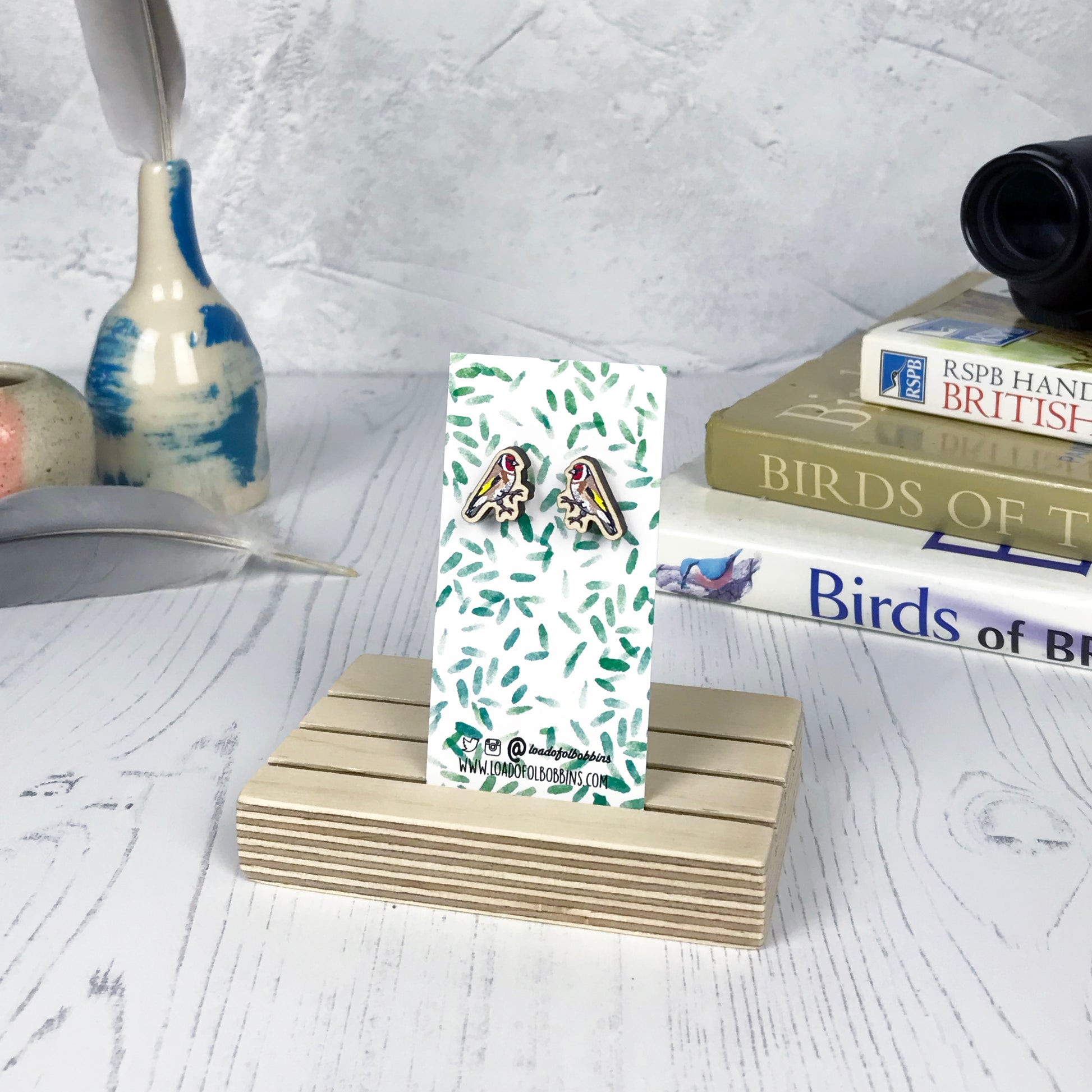 A pair of laser cut wooden earrings showing an illustration of a goldfinch bird by loadofolbobbins. The earrings are mounted on a mini backing card with a repeat abstract watercolour leaf pattern. They are standing in a wooden stand and in the background you can see a pile of bird watching books and a pair of binoculars.