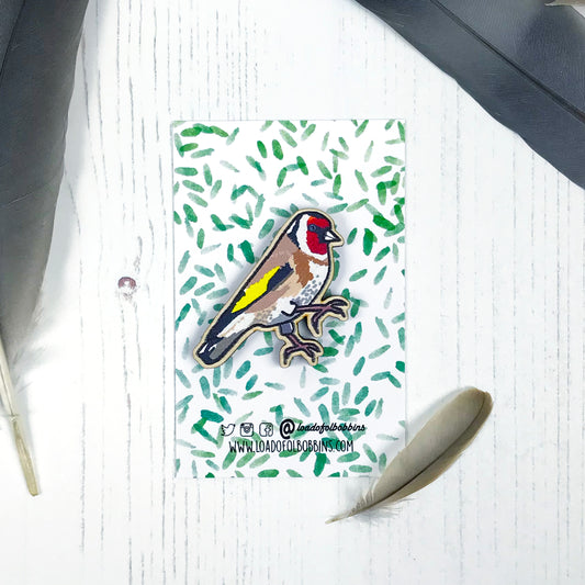A laser cut wooden brooch showing an illustration by Loadofolbobbins of a goldfinch. The brooch is mounted on a backing card with a repeat abstract watercolour leaf pattern.