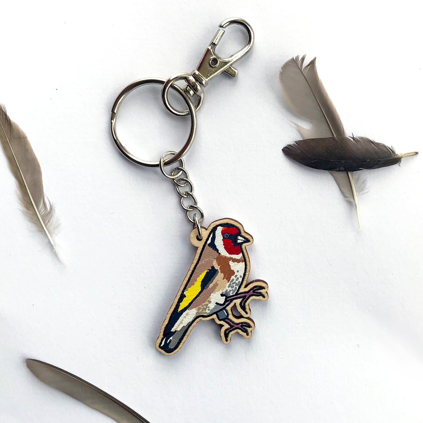 A laser cut wooden key ring showing an illustration of a goldfinch bird by loadofolbobbins. The wooden charm is hanging from a silver colour metal chain, key ring and bag clasp.