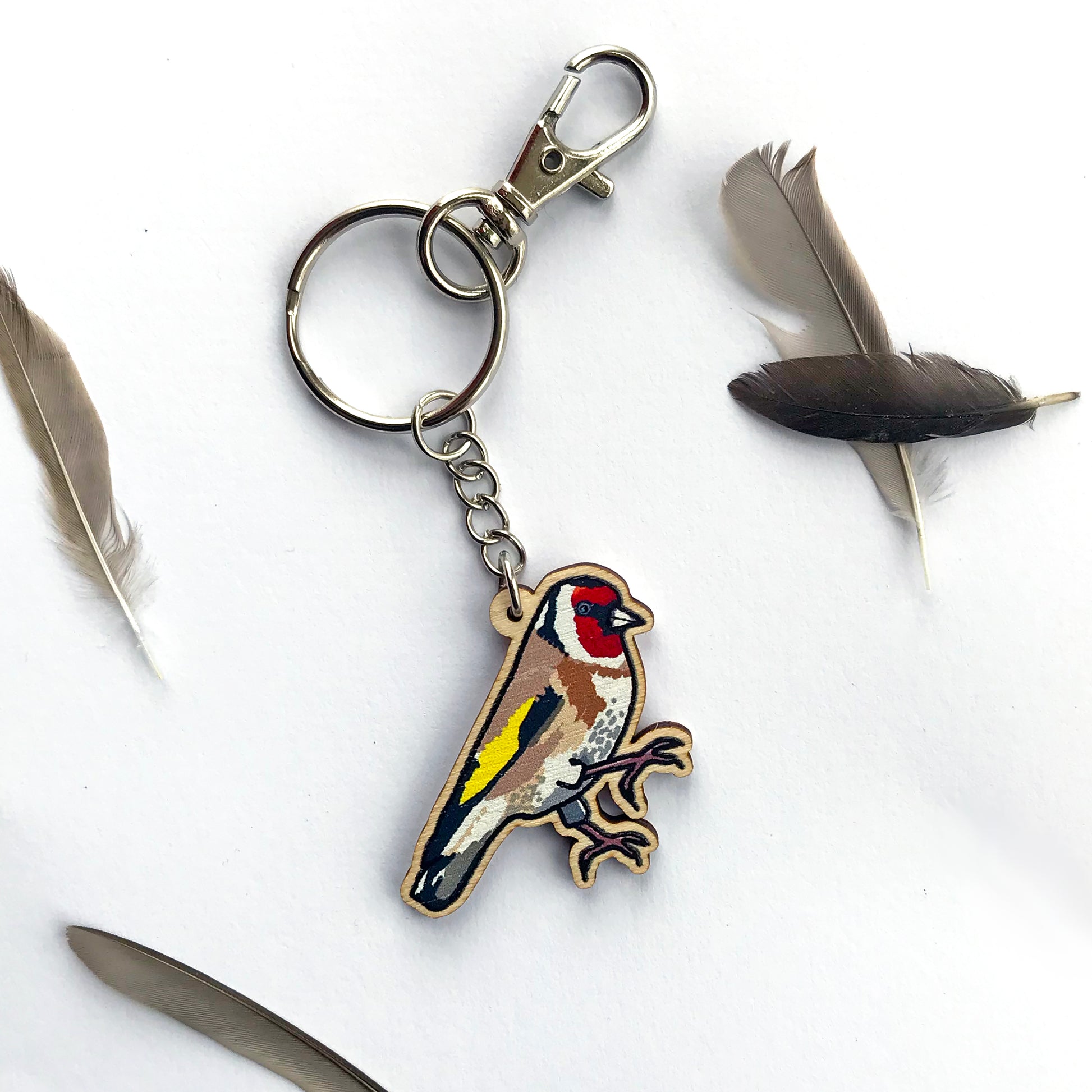 A laser cut wooden key ring showing an illustration of a goldfinch bird by loadofolbobbins. The wooden charm is hanging from a silver colour metal chain, key ring and bag clasp.