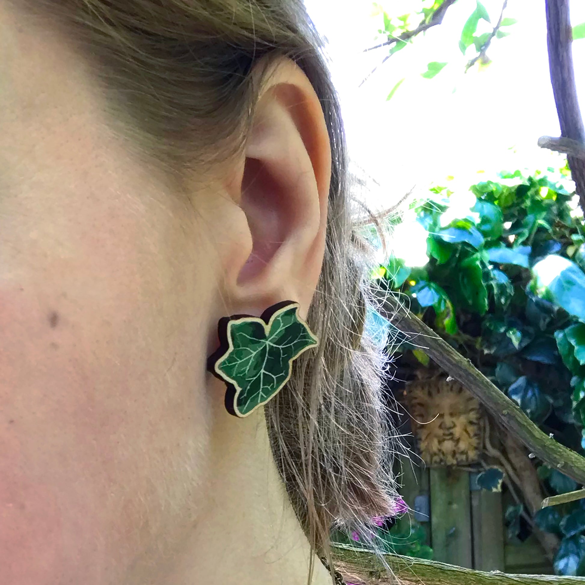 A close up photo of a white woman's ear with blondish brown hair worn up and standing against a wall of ivy. The main focus of the image is her ear which is wearing a small laser cut wooden stud earring of a dark green ivy leaf by Loadofolbobbins.