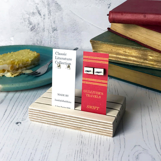 Laser cut wooden stud earrings by loadofolbobbins showing a silhouette scene inspired by the book Gulliver's Travels by Johnathan Swift. It shows Gulliver being tied to the ground by lilliputians. The scene is printed against original text from the novel. The earrings are mounted on a red backing card with gold lettering detail and made to look like the spine of a book.