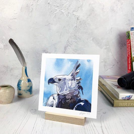 A photo of a small sized 14 cm square art print made from an original illustration by Loadofolbobbins. It is showing a side profile of the head and upper wings of a harpy eagle. The feathers at the back of it’s head are standing upright and proud. The feathers are greys, browns and greyish blues and it has a powerful looking curved pointed beak. The illustration background is a blue inky wash.. There is a 2.5 cm white border for framing.
