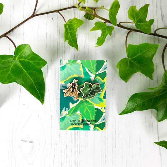 A pair of laser cut mix and match wooden stud earrings showing illustrations by Loadofolbobbins of a dark green ivy leaf and a golden hoverfly. The earrings are mounted on a backing card with a repeat ivy leaf pattern.