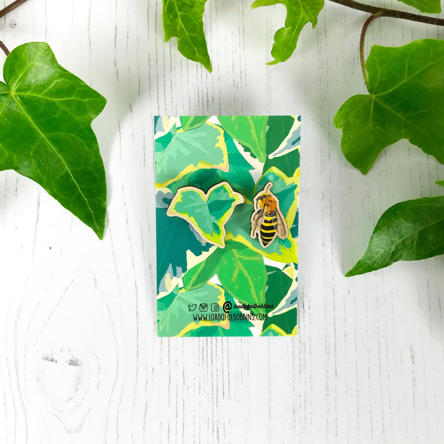 A pair of laser cut mix and match wooden stud earrings showing illustrations by Loadofolbobbins of a light green ivy leaf and an ivy mining bee. The earrings are mounted on a backing card with a repeat ivy leaf pattern.