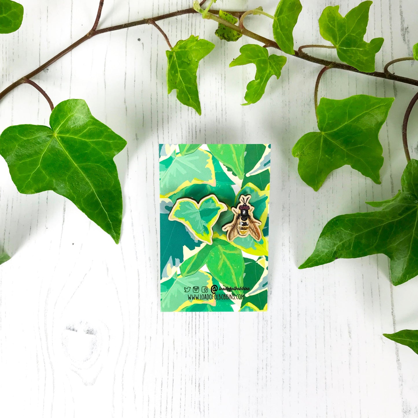 A pair of laser cut mix and match wooden stud earrings showing illustrations by Loadofolbobbins of a light green ivy leaf and a golden hoverfly. The earrings are mounted on a backing card with a repeat ivy leaf pattern.